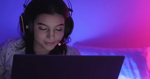 Woman gamer in bed. Caucasian woman on bed playing multiplayer game on laptop at night with color contrast. Happy winning mood. Front shot with copy space.