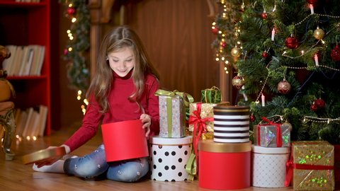 Cute girl at night sitting on the floor near the Christmas tree opens gifts and has fun.