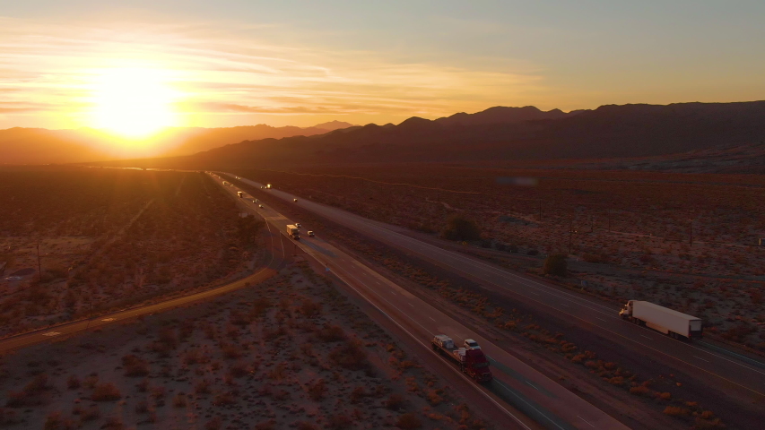 AERIAL, LENS FLARE: Scenic shot of 18 wheeler trucks and cars crossing Mojave desert at dusk. Golden evening sun rays shine on the traffic moving up and down the straight freeway in rural California. | Shutterstock HD Video #1042216279
