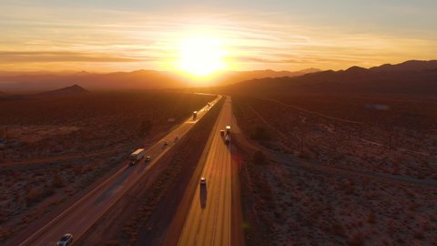 AERIAL, LENS FLARE: Scenic shot of 18 wheeler trucks and cars crossing Mojave desert at dusk. Golden evening sun rays shine on the traffic moving up and down the straight freeway in rural California.