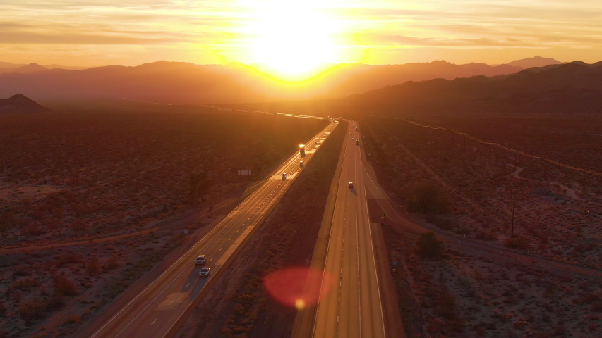 AERIAL, LENS FLARE: Flying above trucks and cars crossing the Mojave desert at sunset. Freight trucks and cars move along the asphalt highway running through the California wilderness on sunny evening Royalty-Free Stock Footage #1042216282
