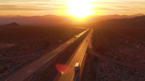 AERIAL, LENS FLARE: Flying above trucks and cars crossing the Mojave desert at sunset. Freight trucks and cars move along the asphalt highway running through the California wilderness on sunny evening
