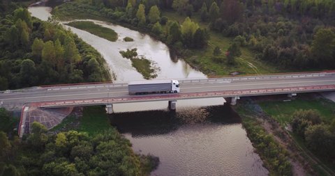 One White Truck with trailer driving on bridge through mountain river / Freeway traffic at summer sunset in hilly landscapes - Aerial drone view
