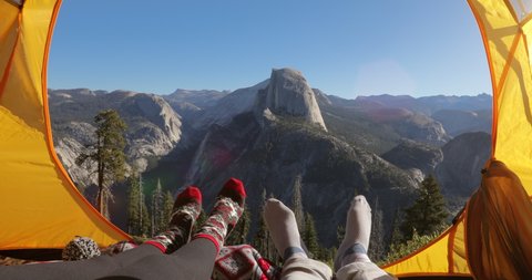 Camping rest in Yosemite Valley. A couple lay inside the yellow tent and point their fingers at the famous Half Dome rock that is seen outside on a picturesque mountain ridge. Yosemite National Park