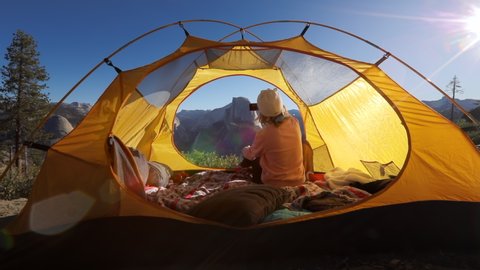 The girl shoots scenic views of the Yosemite Valley, Sierra Nevada ridge and the Half Dome cliff on a smartphone. She sits in a tourist tent flooded with the rays of rising sun. USA.