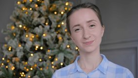 Video portrait of young positive hipster, student or entrepreneur woman in plaid shirt smiling and looking at camera in room with Christmas tree. Garland light illumination. People and holiday concept