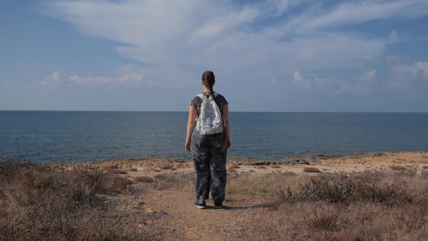 Young girl or woman stands at coastline and looks at autumn sea, rear view