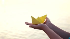 Happy travelling concept. Cute white kid standing at morning sunny golden beach holding paper folded boat in hands. Real time 4k video footage.