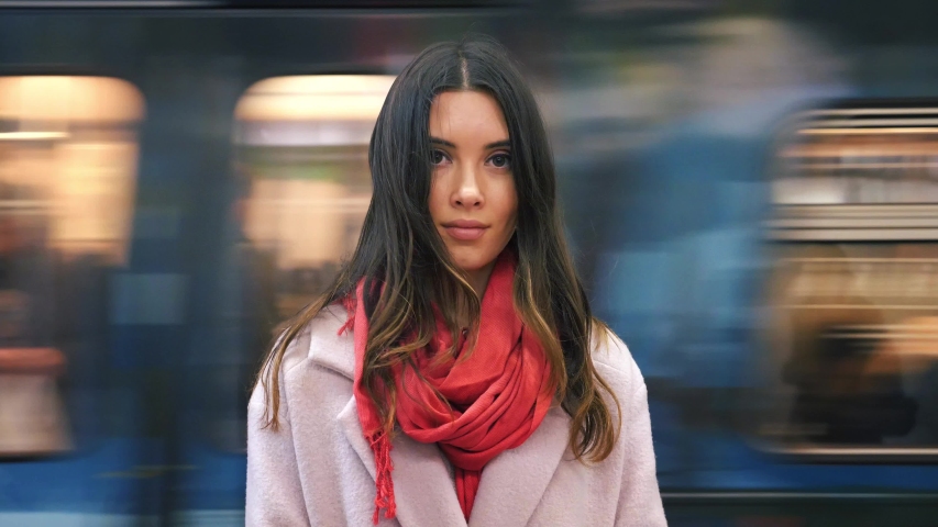 Young woman in red scarf smiling, looking at camera. Blurred subway train moving in background. Slow motion