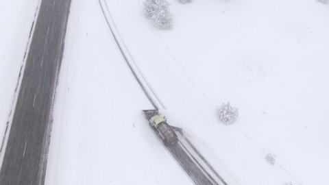 AERIAL: Cars drive past a snow ploughing truck clears and salts a highway lane during an intense snowstorm. Flying behind a snowplow removing fresh snow gathered on the side of an asphalt freeway.