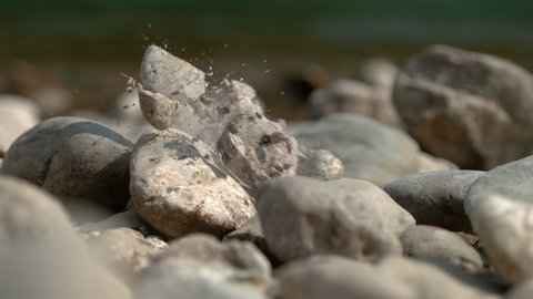 SLOW MOTION, MACRO, DOF: Stone breaks and shatters into pieces after falling onto the riverbank. Cinematic shot of stone breaking into pieces. Fragments of a large grey pebble fly around the shore.