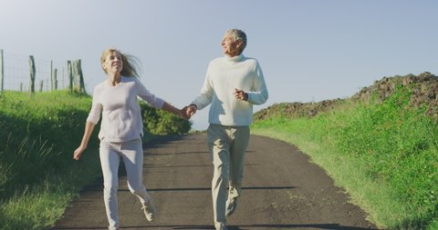 Happy retired couple having fun frolicking together on a country road, older couple skipping and running happily outdoors together on a sunny day with green fields smiling couple enjoying retirement