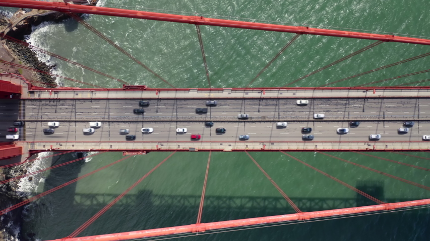 Many vehicles drive along the Golden Gate Bridge road. Bridge roadbed goes from above the water and to the land. Aerial topdown footage. Famous tourist attraction of San Francisco, California. 4k Royalty-Free Stock Footage #1042299577