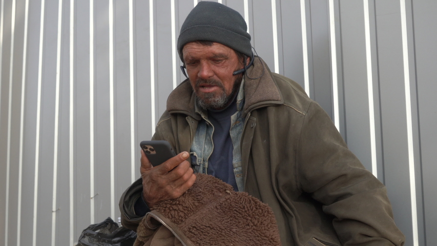 Homeless man listening to music with earphones and mobile phone singing along with the song on smart phone. 50 year old wretched beggar living on the streets for 20 years.