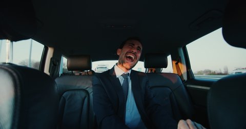 An young handsome businessman rejoices and celebrates success in the financial and economic fields during traveling in modern car with a driver in center of the city by day with a sunshine. Shot in 8K