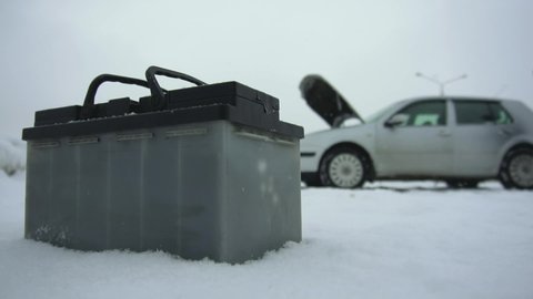 Man repairs or fixes broken car in winter snowfall. Malfunction or problem with auto. Automotive battery lying next to the vehicle. Hood or bonnet raised up. Typical lead-acid battery is on foreground
