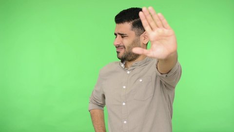 Dissatisfied brunette man with bristle in shirt showing stop sign with outstretched hand and saying no, don't show me this, deny reject concept. indoor studio shot, green background, chroma key