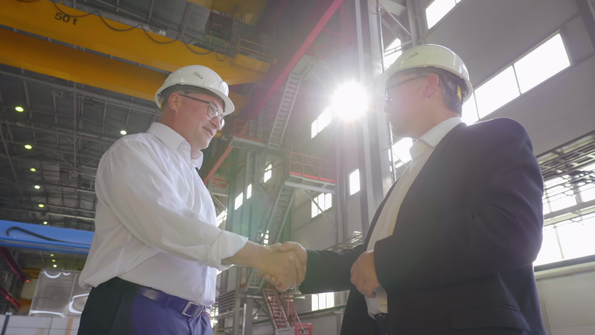 Slow motion shot of asian and caucasian engineers or businessmen shake hands in factory workshop. Successful deal after negotiations. Handshake of business people in hard hats in plant facility