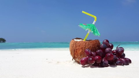 Open fresh hairy coconut with the cocktail and straw decorated with umbrella and grape on the white sand beach. Ibiza party concept