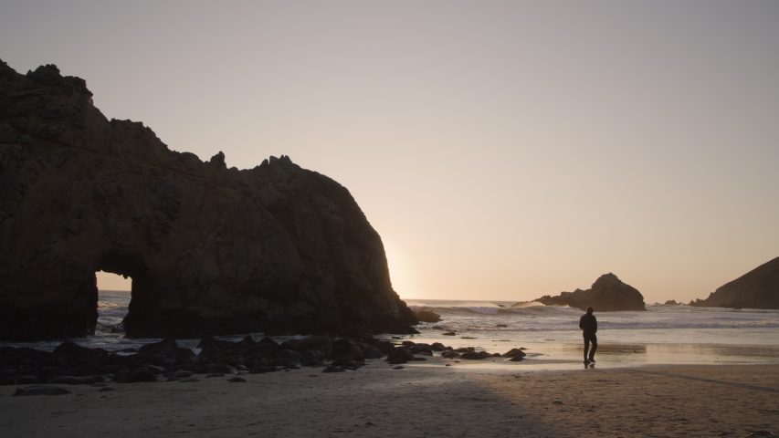Wide panning shot of distant man walking near Keyhole Arch at sunset / Big Sur, California, United States