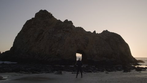 Wide shot of distant woman photographing Keyhole Arch at sunset / Big Sur, California, United States