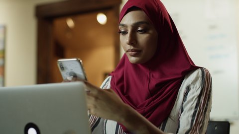 Close up of serious woman wearing hijab texting on cell phone then typing on laptop / Cedar Hills, Utah, United States