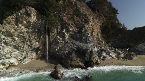Aerial view zooming out from waves splashing on rocks in ocean near waterfall / Big Sur, California, United States