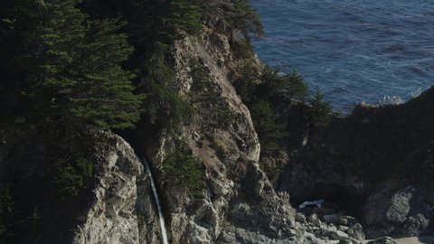 Tilt down to high angle view of waves splashing on rocks in ocean near waterfall / Big Sur, California, United States
