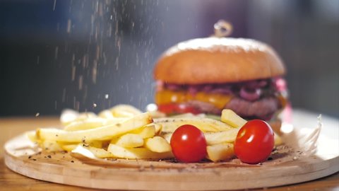 Crispy french fries with hamburger on wooden board. Black pepper sprinkles on french fries and burger in slow motion. Fast food concept.