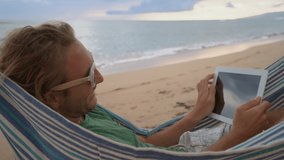 Young man using digital touch screen tablet reading and enjoying free time on an hammock by the beach 
