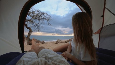 Couple lying down inside a camp tent on the edge of a beautiful beach. Woman looking out at the sunset. Couple camping on deserted island 