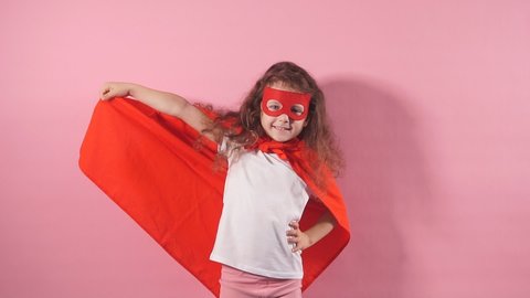 Portrait of confident smiling kid superhero wearing red cloak and mask on eyes imagines herself as hero, stand isolated on pink background