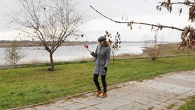 Girl-blogger goes on Park and conducts online broadcast on phone, waves a hand. Woman in autumn coat in the open air talking live and smiling. Technology and lifestyle concept