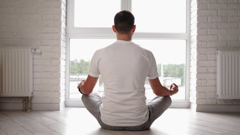 Young modern caucasian man practice yoga lotus pose sitting on the floor in front of the window. Man meditating at home enjoying appeasement and pacification. Harmony wellness. Self isolation