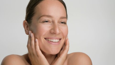 Close-up beauty portrait of young woman with smooth healthy skin, she gently touches her face with her fingers on white background and smiles