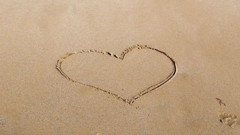 Handwritten heart shape on a sandy sea beach is washed by the sea wave with foam on a summer sunny day. Disappearing love concept