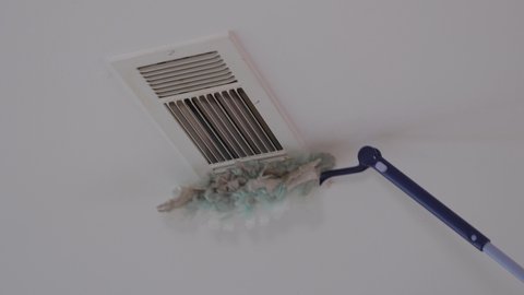 Dusting HVAC ceiling air vent with long handle duster tool. Cleaning overhead home heat and air conditioning ventilation duct.