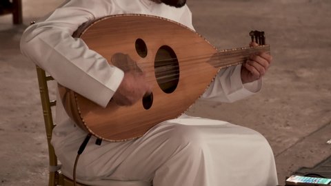 A man in white thobe sat on a chair playing an oud while watching the lyrics from the smartphone placed at front of him.