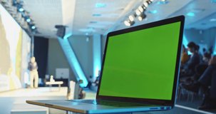 Blank green screen laptop computer in futuristic conference hall with male coach speaking on background. Online education. Chroma key. Business network app working. IT developer. Technology designer.