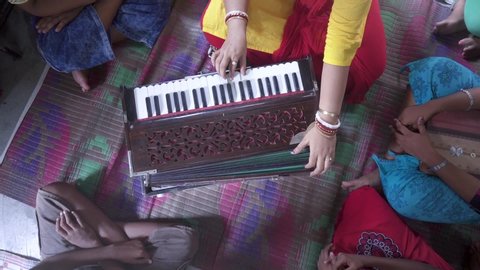 Married Indian teacher teaches music with harmonium at rural classroom with students gathered around, unrecognizable, top down shot