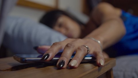 Tired woman in pajamas sleeping in cozy bed, being woken up by alarm clock, reaching out for cellphone on nightstand and snoozing alarm clock on smartphone screen in early morning, unable to wake up.