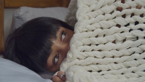 Restless worried african american woman lying awake in bed, covering face with woollen blanket, peeking and looking to the sides in fear. 