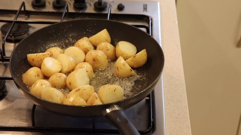 Small potatoes being sautéed in a pan with bubbling butter and garlic on a gas stove. Chopped dried herbs being added and stirred by a woman's hand using a metal spoon