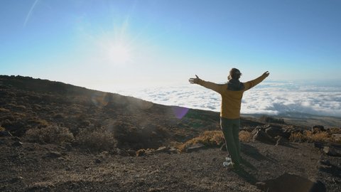 Young man hiker stands above clouds on top of volcano arms wide open to embrace positivity. Contemplative person standing above stunning landscape.