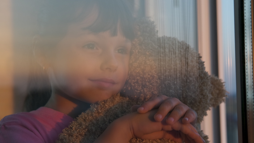 A child with a toy by the window. Portrait of a little girl with a teddy bear looking out the window. Royalty-Free Stock Footage #1042424767