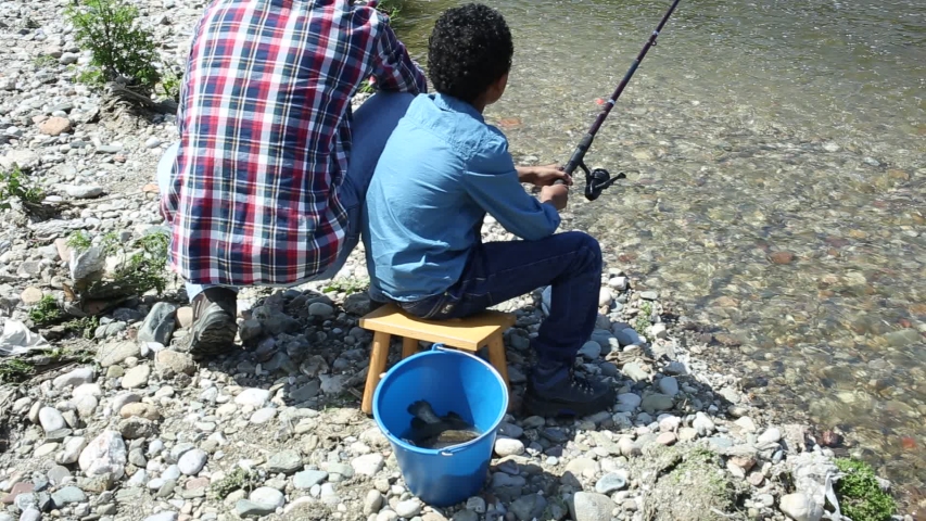 Back portrait of little boy and his father fishing with rods on river
