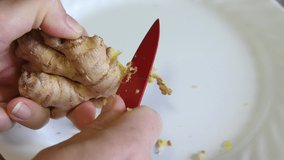 Video peeling skin of ginger root. Removing skin of organic ginger for cooking preparation with red knife. White plate in background. 4K video.