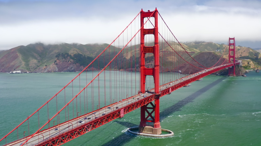 Vibrant aerial 4K footage of the beautiful suspension bridge known as Golden Gate Bridge against the backdrop of the hilly coast and cloudy sky. The water of the strait has turquoise color. Royalty-Free Stock Footage #1042450984