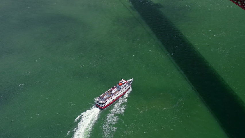 Aerial view over the passenger ship sailing on bright green water. The vessel floats under the famous Golden Gate Bridge, crossing its shadow on the water. San Francisco, California. 4K
