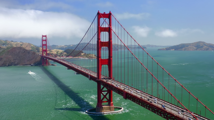 Impressive aerial 4K view over the stately and beautiful Golden Gate Bridge on cloudy summer day. Numerous vehicles move by the bridge in both directions. Calm turquoise water is around